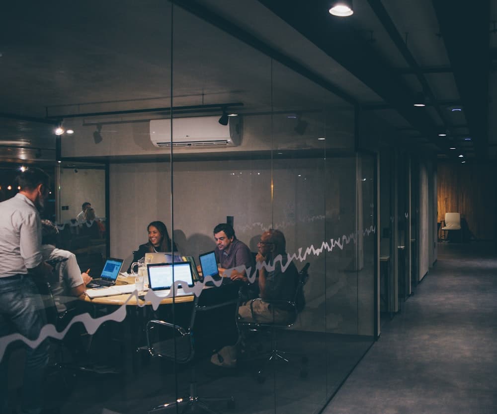 Colleagues working together around a laptop in a glass-walled meeting room.